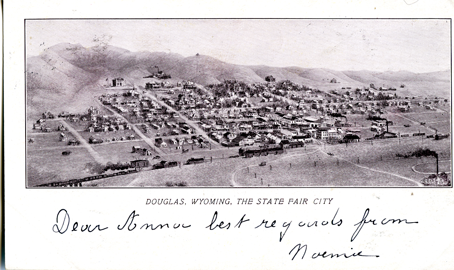 A postcard featuring a pen and ink sketch of a birds-eye view of the tonw of Douglas Wyoming. Small buildings and a grid pattern of of roads are nestled between a train track in the lower front, and a low mountain at the top right corner.